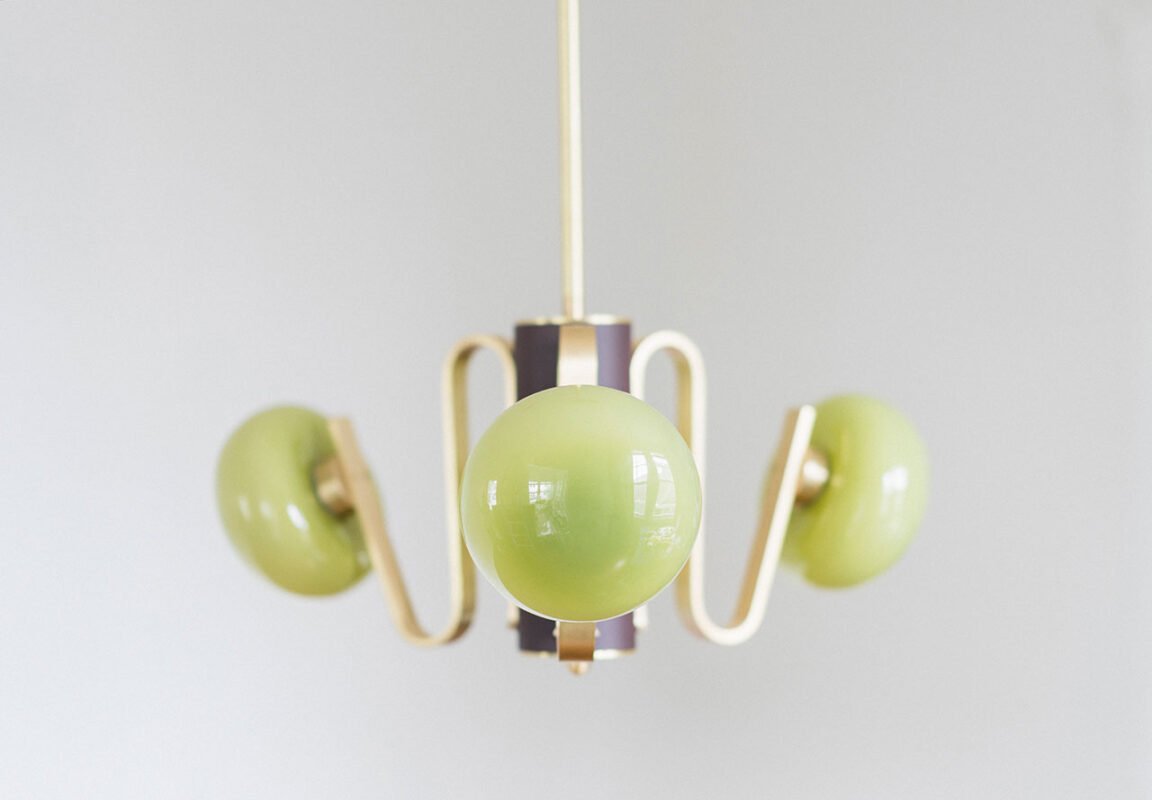 A modern chandelier by Sara Schoenberger, featuring three spherical green glass shades and gold metal arms, suspended against a plain light gray background.