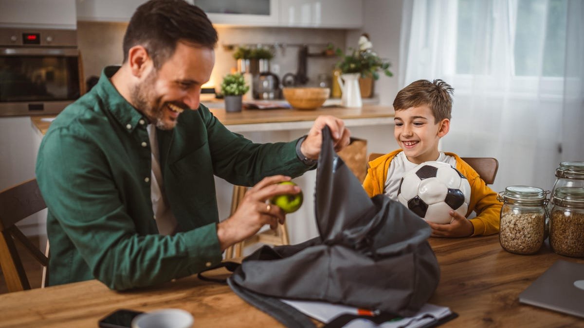 A man and young boy smile as they pack a backpack on a table.