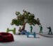 A person sits under a large artificial tree, surrounded by oversized green toy soldiers, a red polka-dotted seat, and two colorful abstract stools in a minimal studio setting.