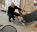 A man wearing glasses and black clothes examines a modern black reclining chair with a metal base in a design studio filled with chairs and sketches.