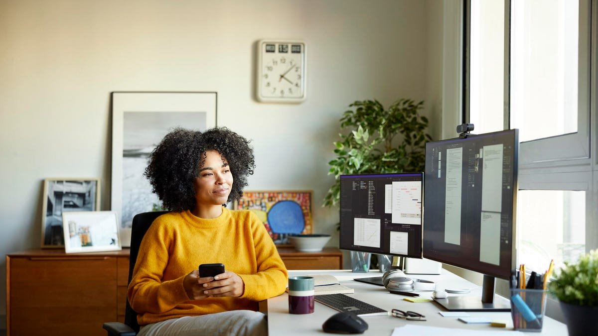 A woman leans back in front of a home desk with a computer and electronics.