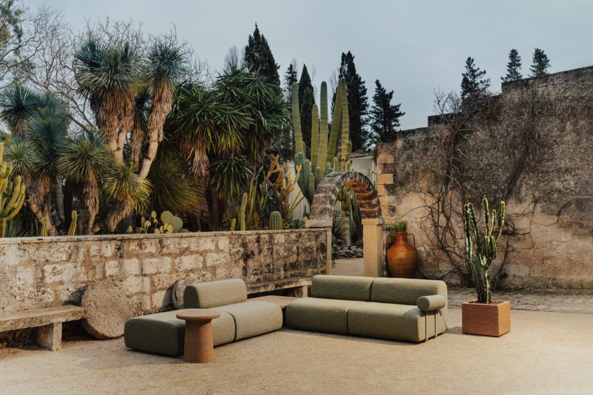 Outdoor seating area with modular sofas, a side table, and potted cacti, set against a stone wall and surrounded by various desert plants and trees.