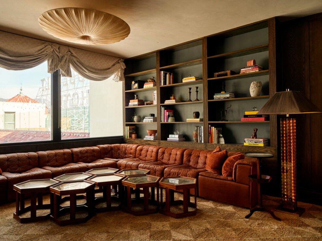 A cozy living room with a brown leather sectional, hexagonal coffee tables, built-in shelves with books and decor, and a large window with draped curtains.